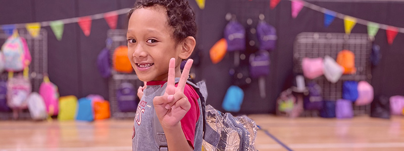 A young boy gives the peace sign while wearing a backpack full of school supplies provided to him and his family